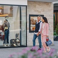 Paar spaziert an einem Modegeschäft in der Region Seefeld vorbei. Auslage mit eleganter Kleidung und Dekohase sichtbar.