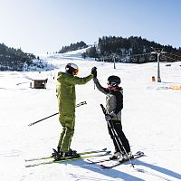 Zwei Skifahrer auf einer verschneiten Piste in der Region Seefeld, die sich einen High-Five geben. Im Hintergrund sind Bäume und ein Skilift.