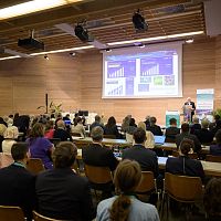 Konferenz mit vielen Teilnehmern und Präsentation auf großen Bildschirmen.