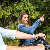 Frau im Rollstuhl zeigt lächelnd in eine Richtung, Mann folgt ihr aufmerksam im Wald.