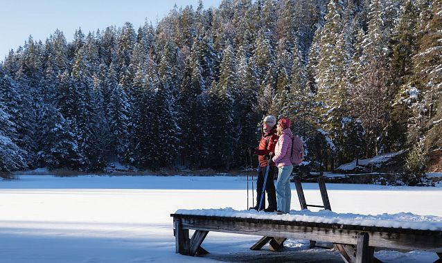 Zwei Personen stehen auf einem Holzsteg am Möserer See, umgeben von schneebedeckten Bäumen. Winterliche Berglandschaft.