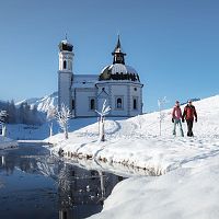 Verschneite Kirche in Winterlandschaft, Wanderer im Vordergrund. Region Seefeld. Klarer Himmel, blau.