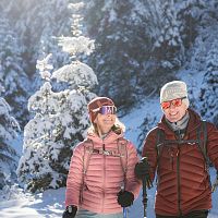 Paar beim Winterwandern im Schnee, mit Jacken und Mützen, umgeben von schneebedeckten Bäumen, in sonniger Winterlandschaft.