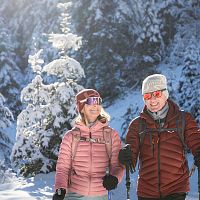 Paar beim Winterwandern im Schnee, mit Jacken und Mützen, umgeben von schneebedeckten Bäumen, in sonniger Winterlandschaft.